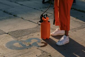 A person in orange pants using an orange sprayer on concrete pavement.